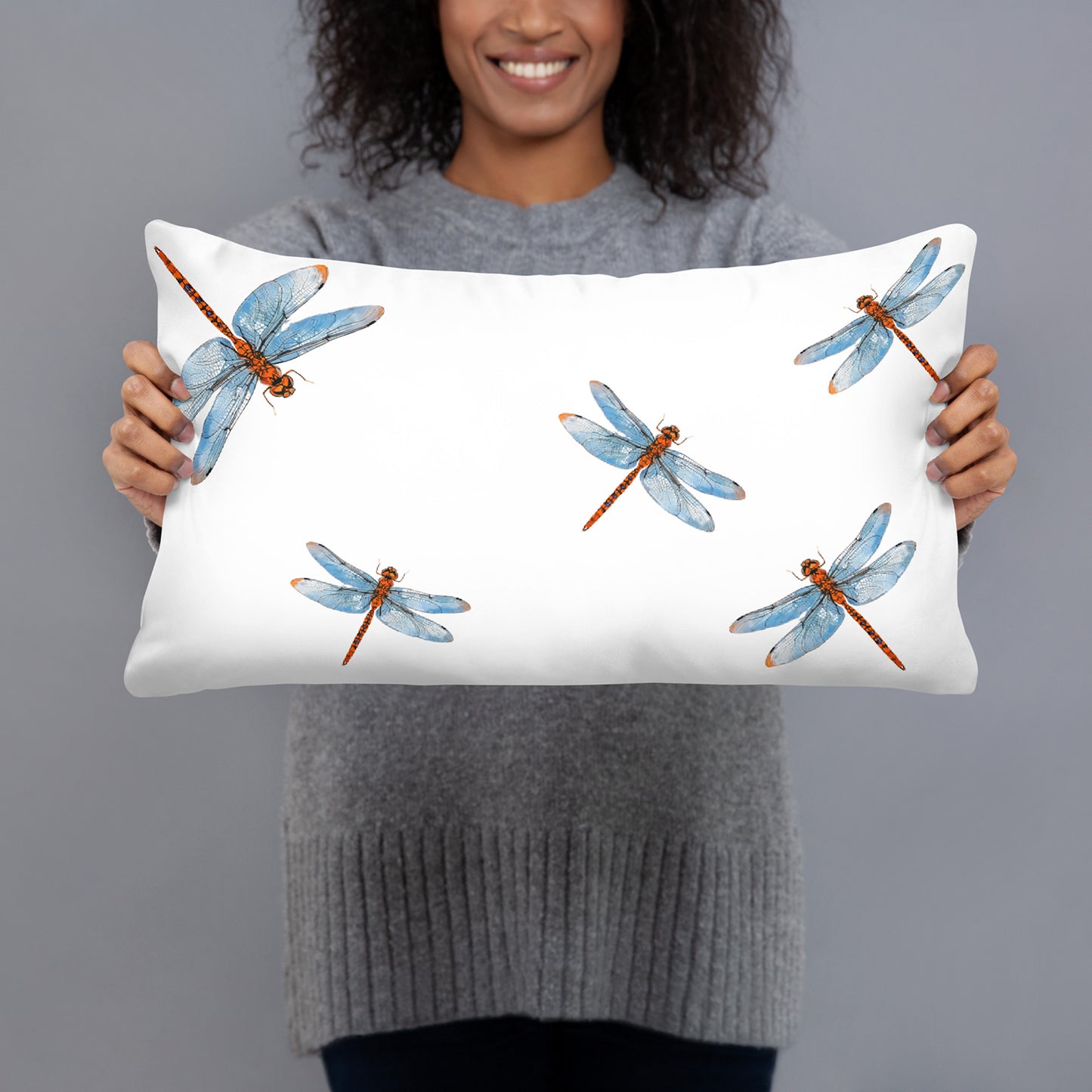 A woman holding a white rectangular lumbar pillow with a watercolor dragonfly pattern. The pillow features five blue and orange dragonflies on a clean white background.