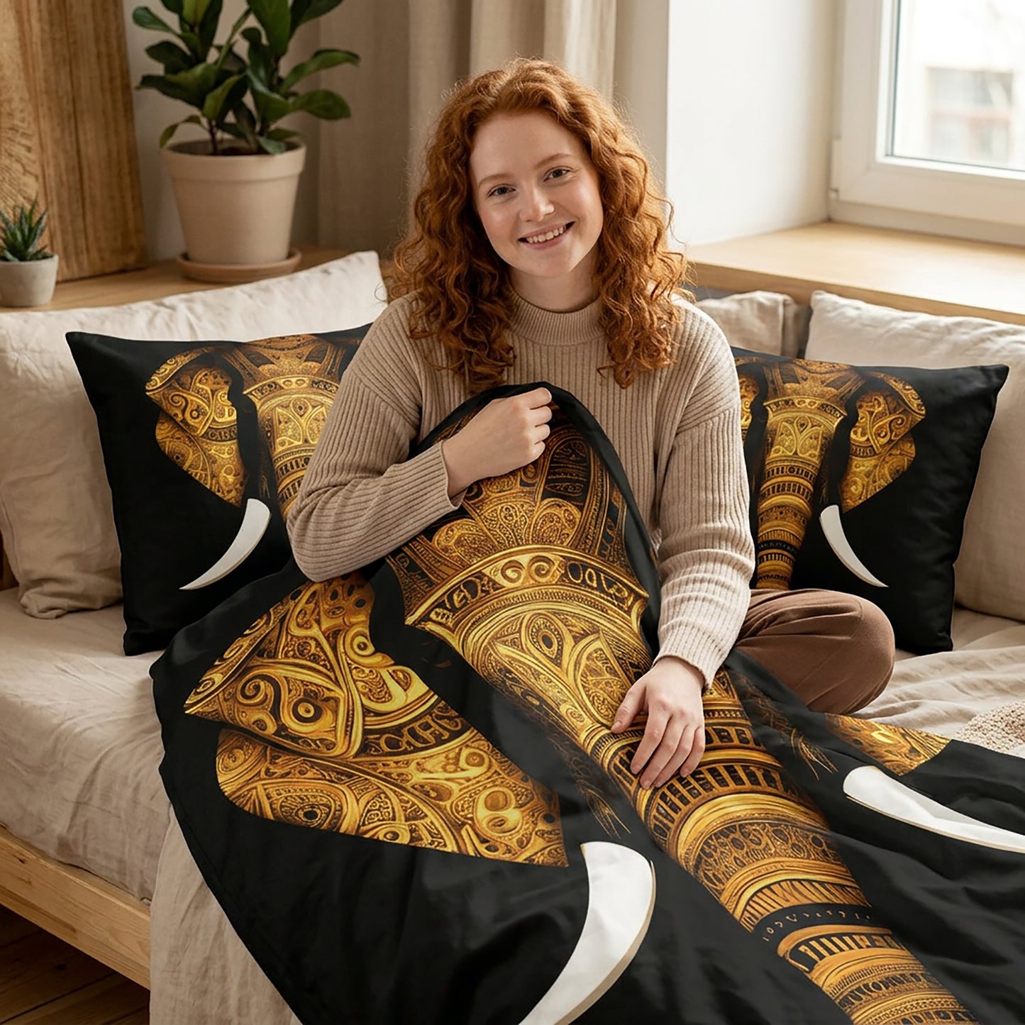A smiling woman with red hair sits on a bed holding a black and gold patterned duvet cover, with matching black pillows featuring gold and white elephant designs in a bright bedroom.