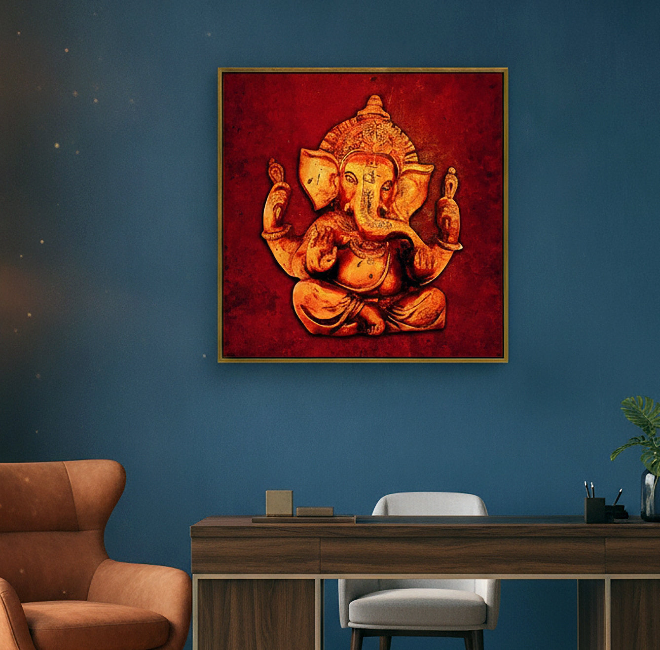 A square framed wall art print featuring a golden Ganesha figure on a textured red background, displayed on a dark blue wall above a wooden desk in a modern room setting.