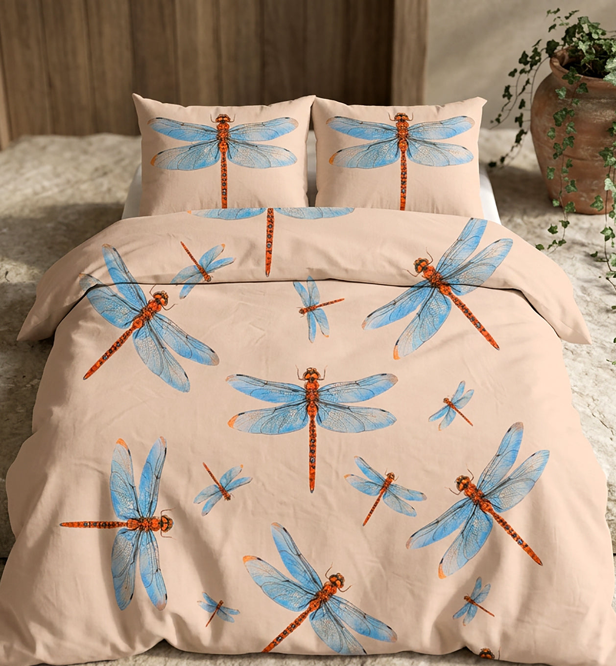 A bedding set featuring a duvet cover and two pillowcases with a beige background and a pattern of blue dragonflies with orange-brown bodies, styled on a bed in a bedroom setting.