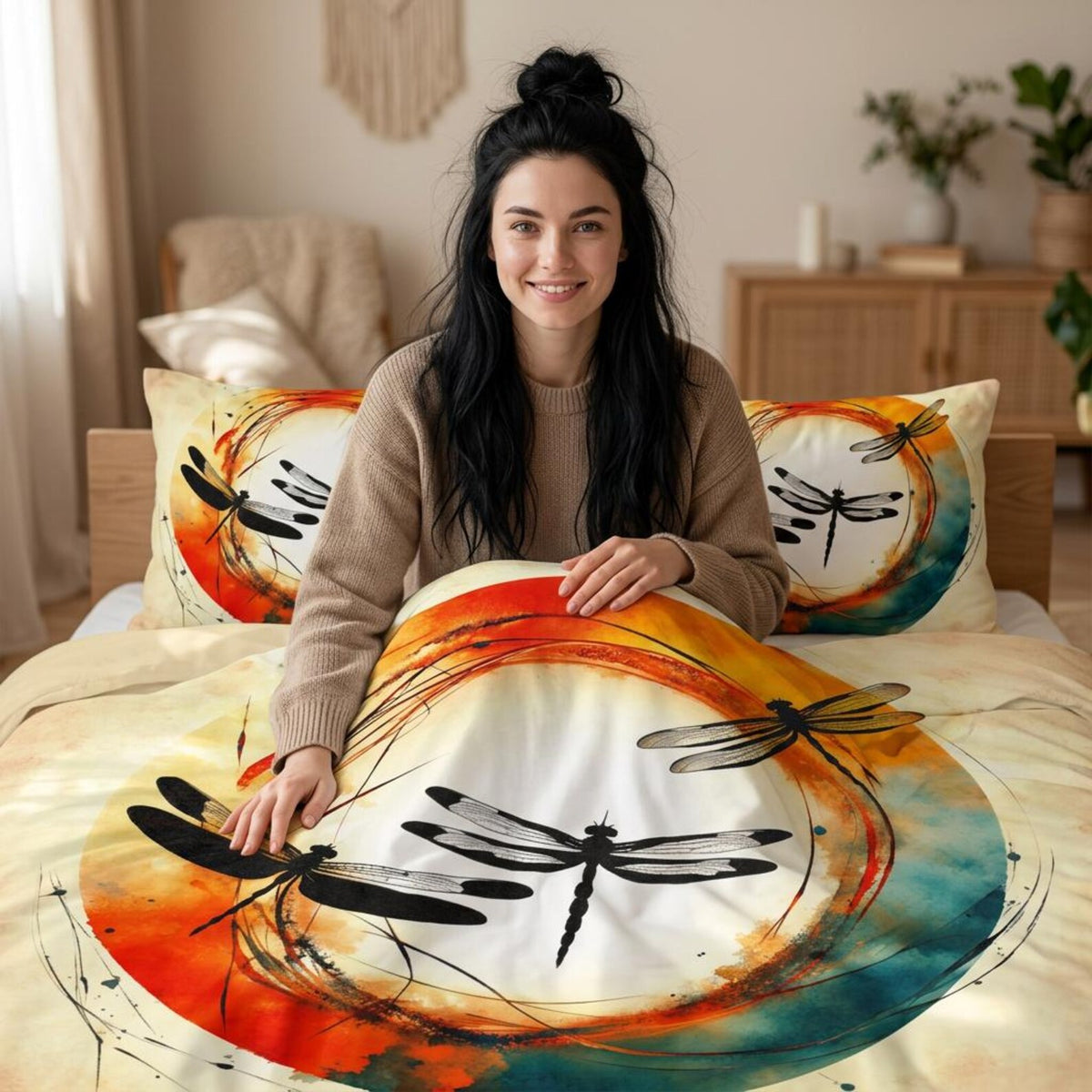 A woman sits on a bed in a warmly lit bedroom, wrapped in a duvet cover and pillows featuring a large dragonfly print over orange, yellow, teal, and cream watercolor-style circles.