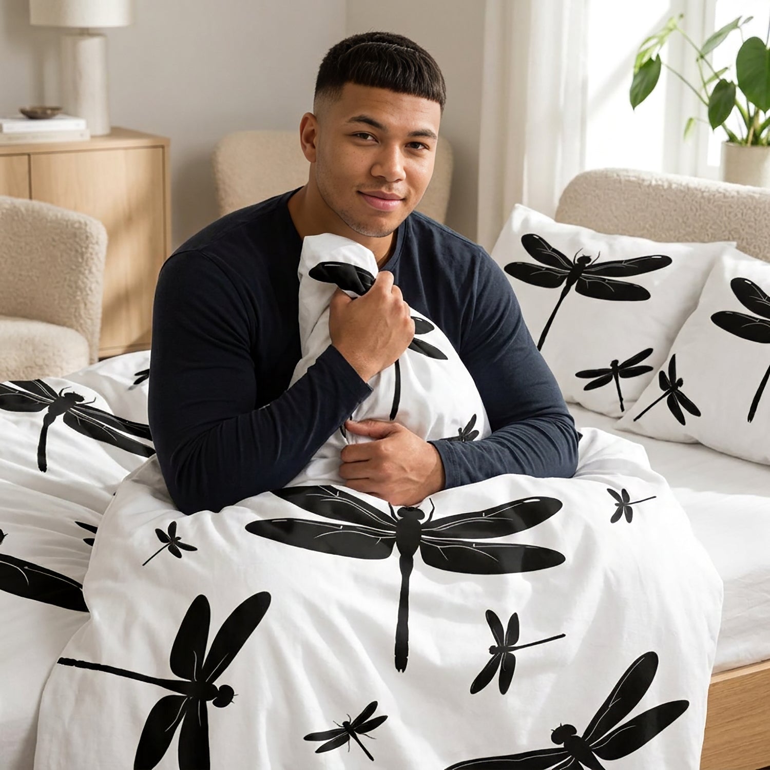 A man sitting on a bed wearing a navy long-sleeve shirt and holding a white pillow with a black dragonfly print, with a matching white duvet featuring black dragonfly patterns spread across the bed in a bright bedroom setting.