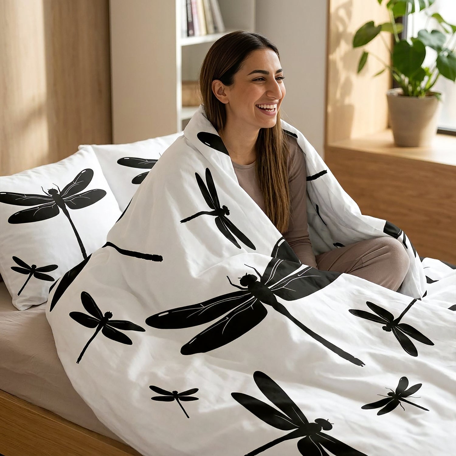 A woman sitting on a bed wrapped in a white duvet cover with a black dragonfly pattern, in a bright, modern bedroom.