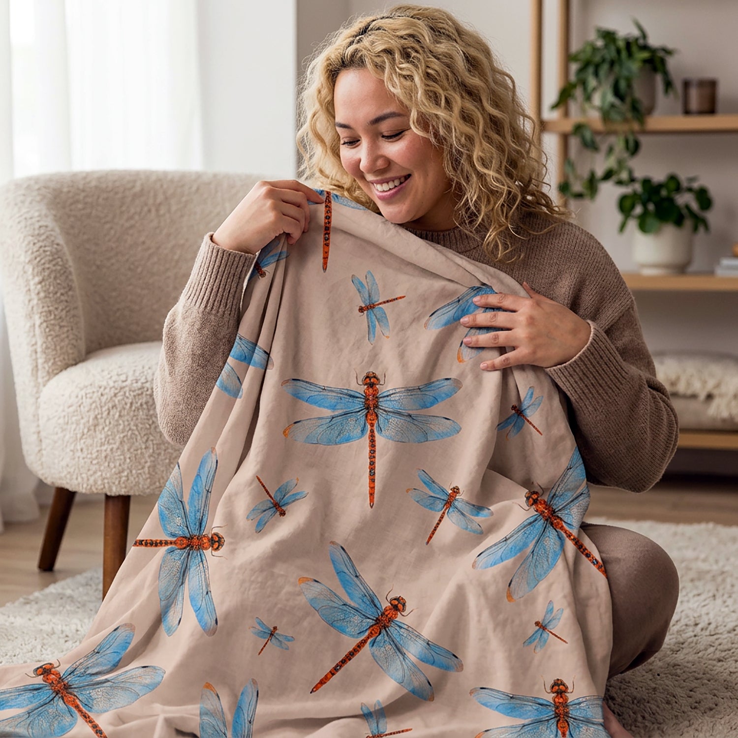 A woman sitting on a rug in a cozy living room setting holds up a beige blanket featuring a pattern of blue dragonflies and orange-red dragonfly bodies.