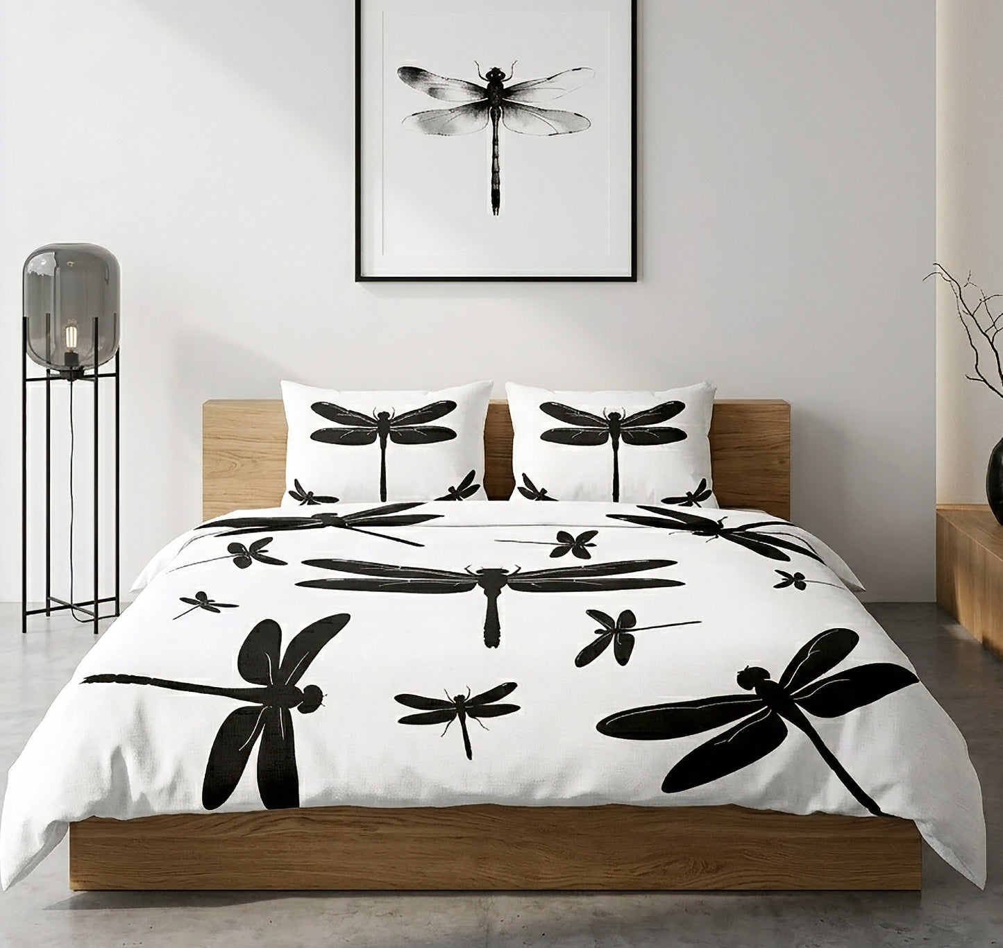 A bed dressed in a white duvet cover and matching pillowcases, printed with large black dragonfly silhouettes, shown in a modern bedroom setting with a wooden bed frame, wall art, and a floor lamp.