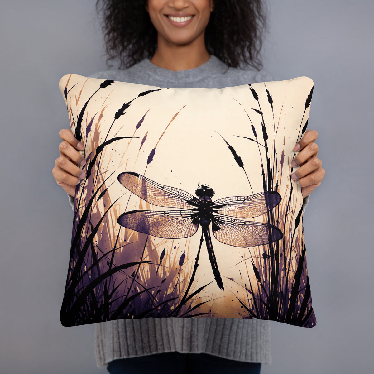 A person holding a square decorative throw pillow featuring a printed dragonfly and tall grass design in black, purple, and cream tones against a plain gray studio background.