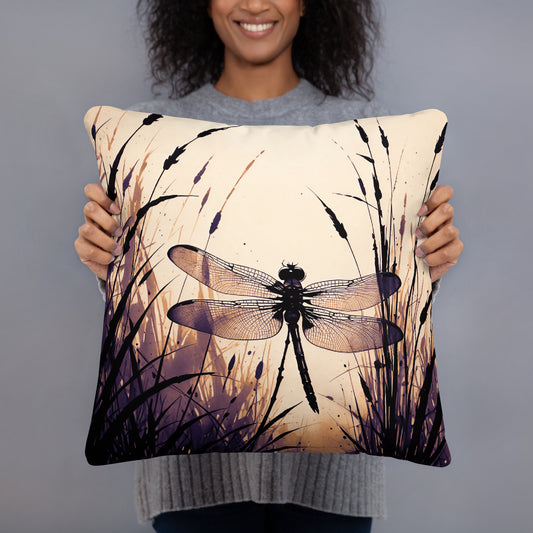 A person holding a square decorative throw pillow featuring a printed dragonfly and tall grass design in black, purple, and cream tones against a plain gray studio background.
