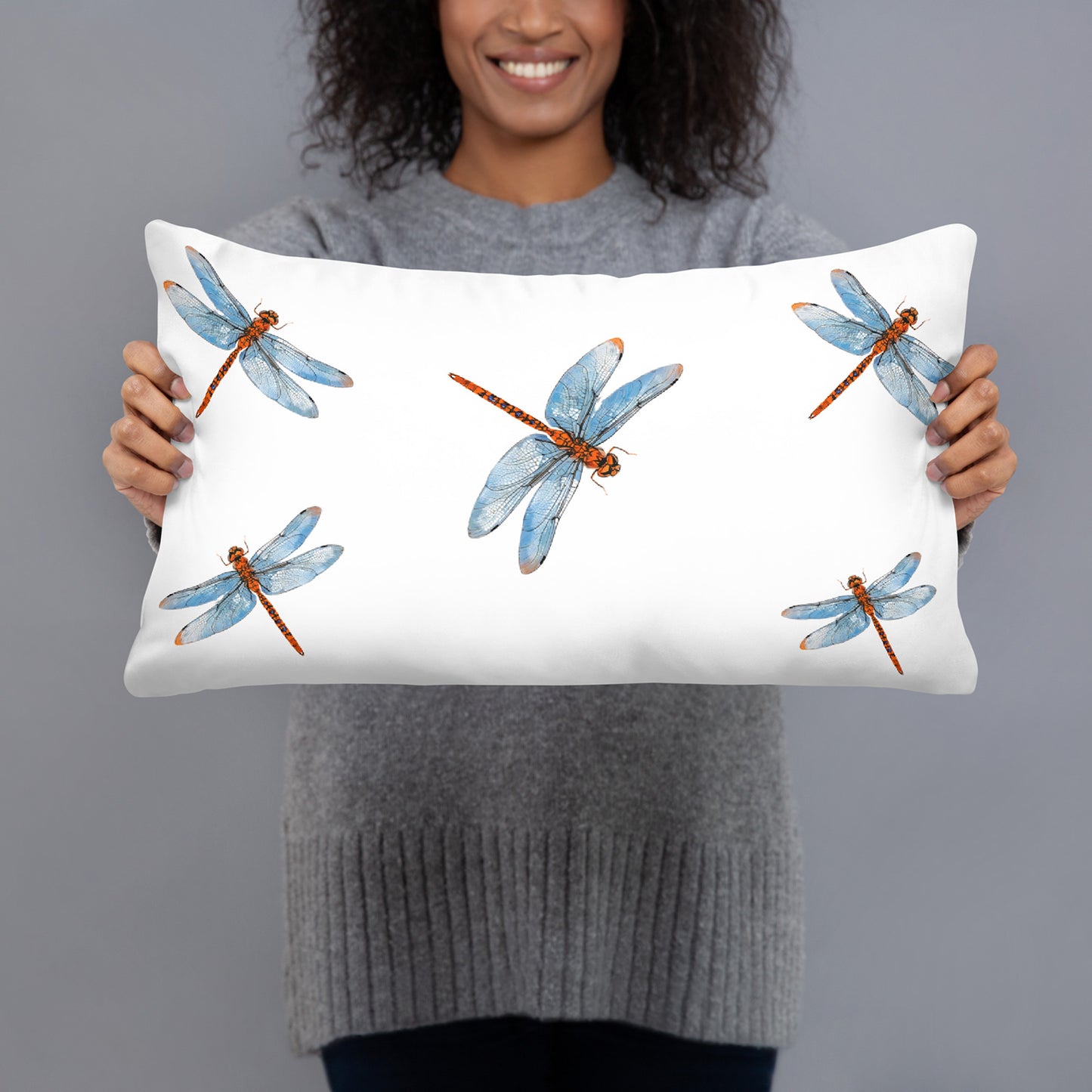 A woman holding a white lumbar pillow with a watercolor dragonfly pattern. The pillow features four blue and orange dragonflies on a white background, and the woman is wearing a gray ribbed sweater against a solid gray studio background.