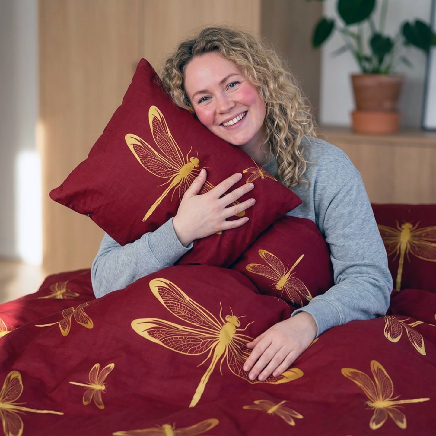 A smiling woman with curly blonde hair sits in bed holding a red pillow and covered with a matching red duvet featuring large yellow dragonfly prints. A wooden headboard and a potted plant are visible in the softly lit room.