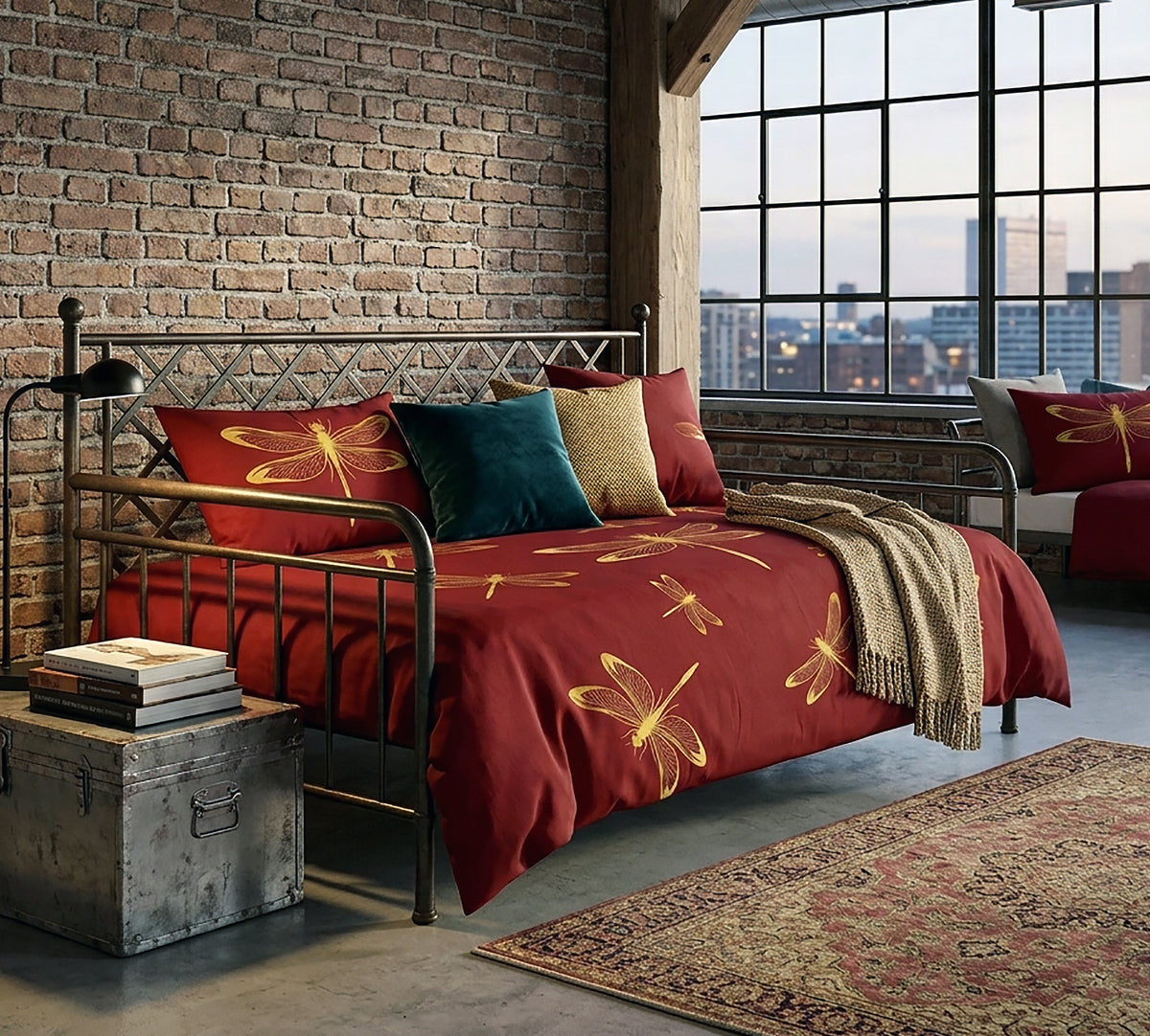 A bed dressed in a red duvet cover and matching pillow shams, decorated with large gold dragonfly prints, styled in a loft bedroom with a brick wall, large window, and city view.