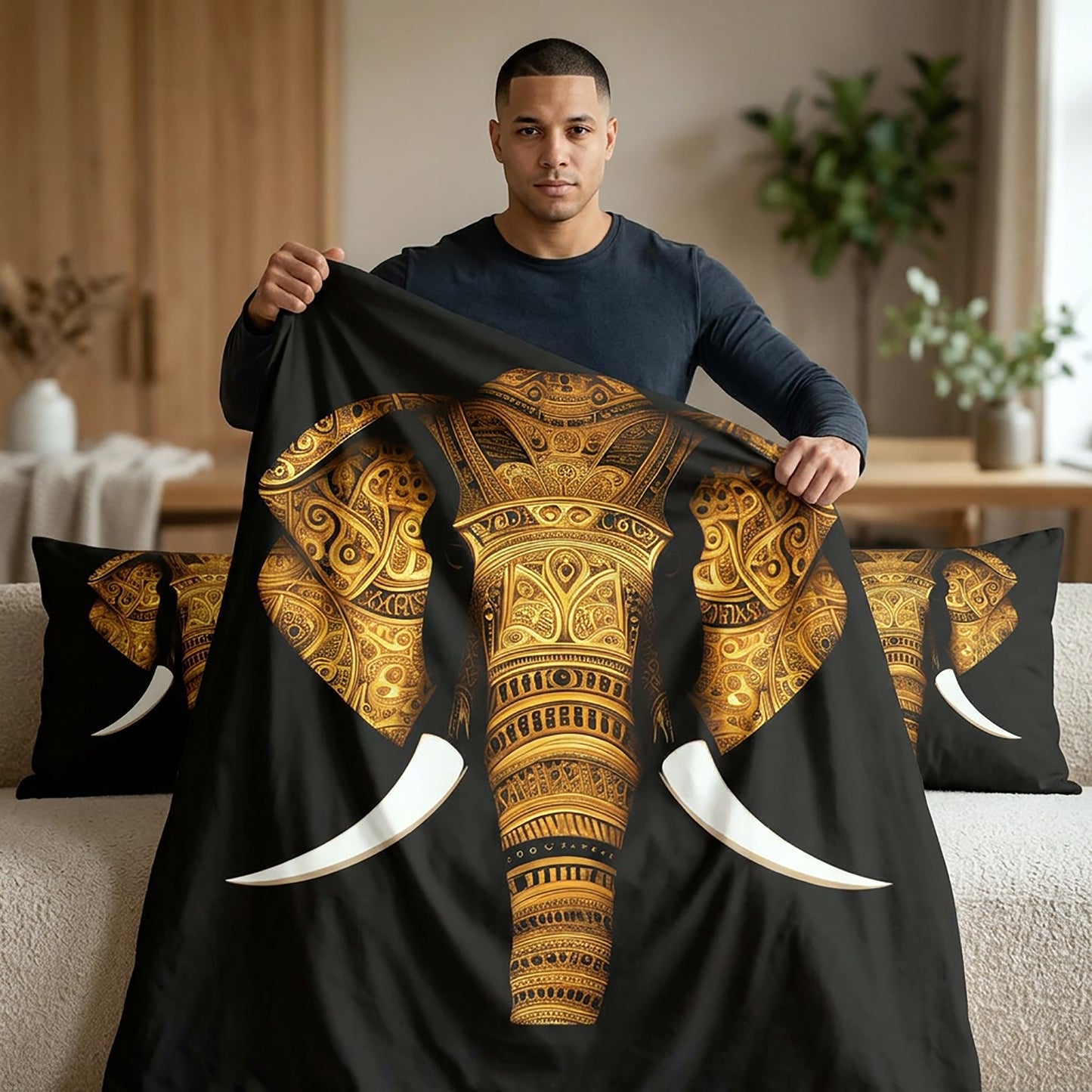 A man holds up a black bedding set featuring a large gold and brown patterned elephant design with white tusks, shown with matching pillowcases in a modern living room setting.