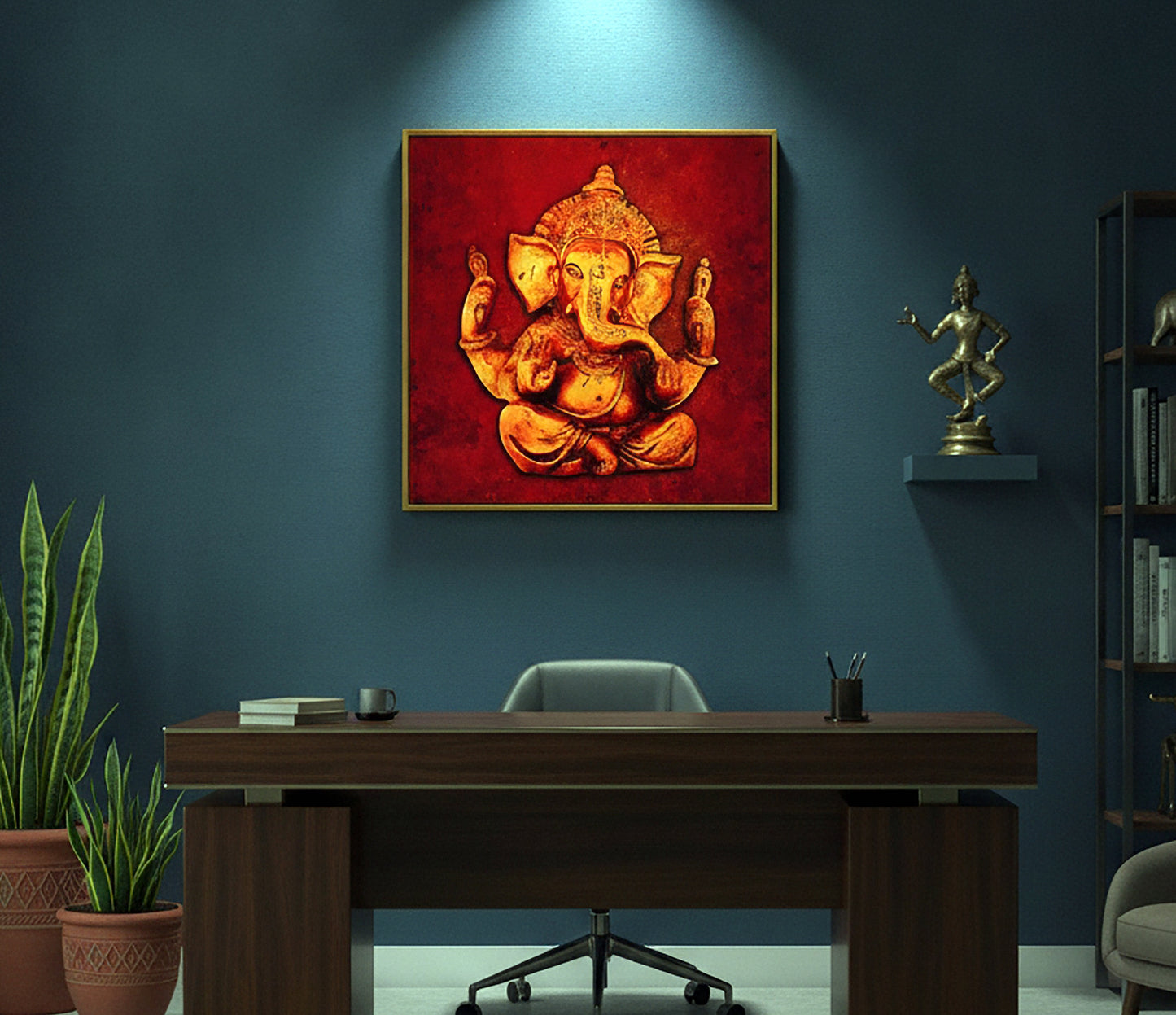 A framed canvas art print of Lord Ganesha in gold and orange tones against a textured red background, displayed on a dark blue wall above a wooden desk in a styled office setting.