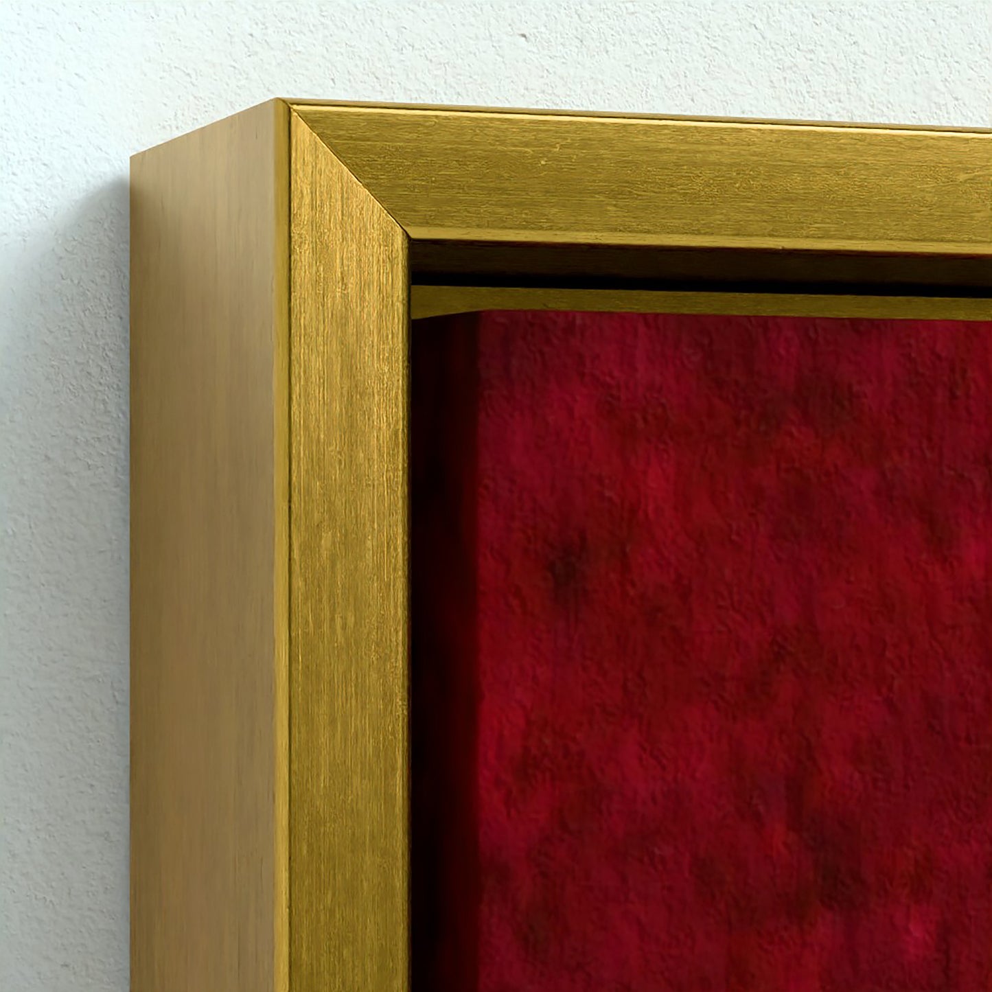 Close-up of a framed wall art piece with a brushed gold frame and a deep red textured fabric or velvet insert against a white wall.