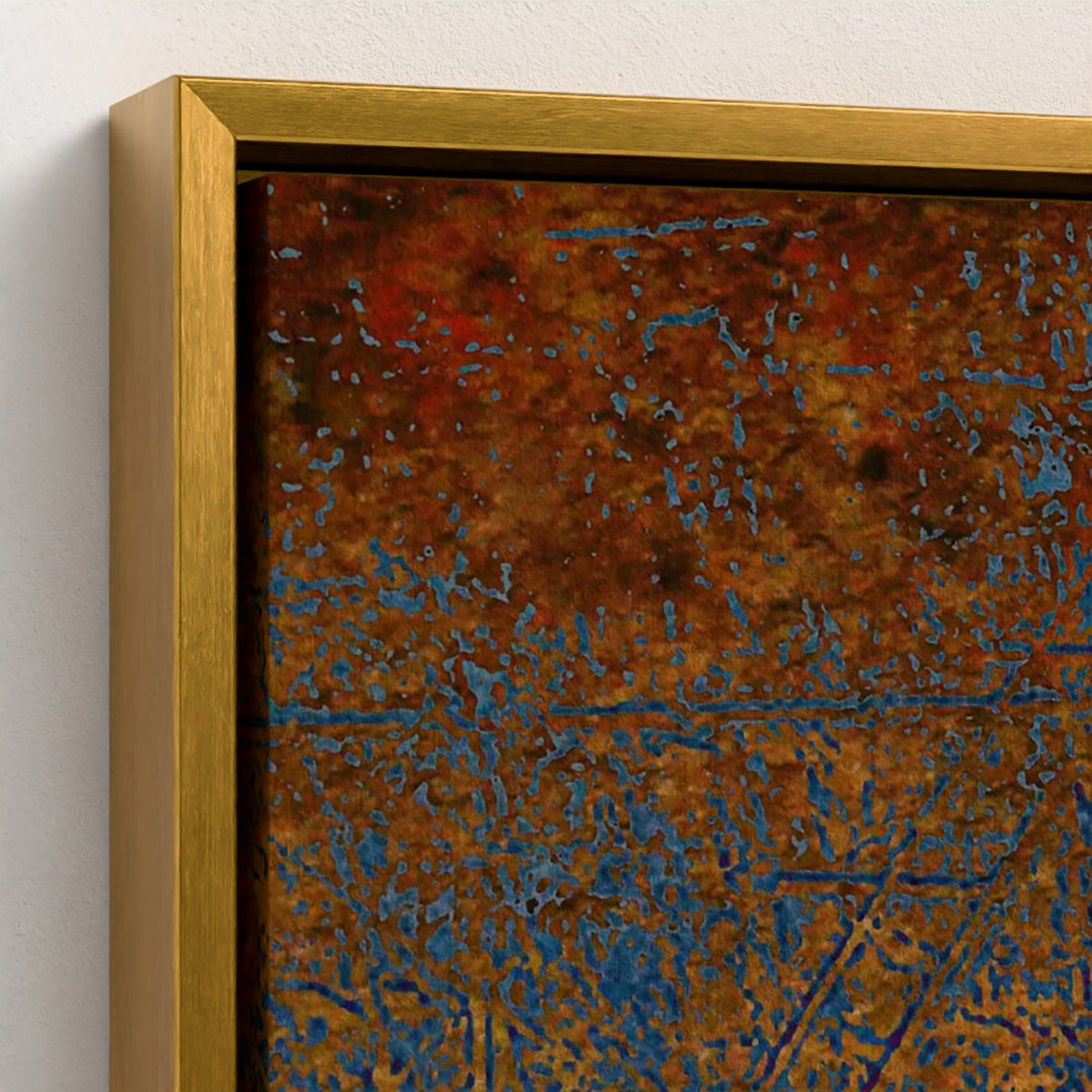 Close-up of a framed abstract artwork with a textured rust-brown and blue surface, shown with a gold-toned frame against a light wall.