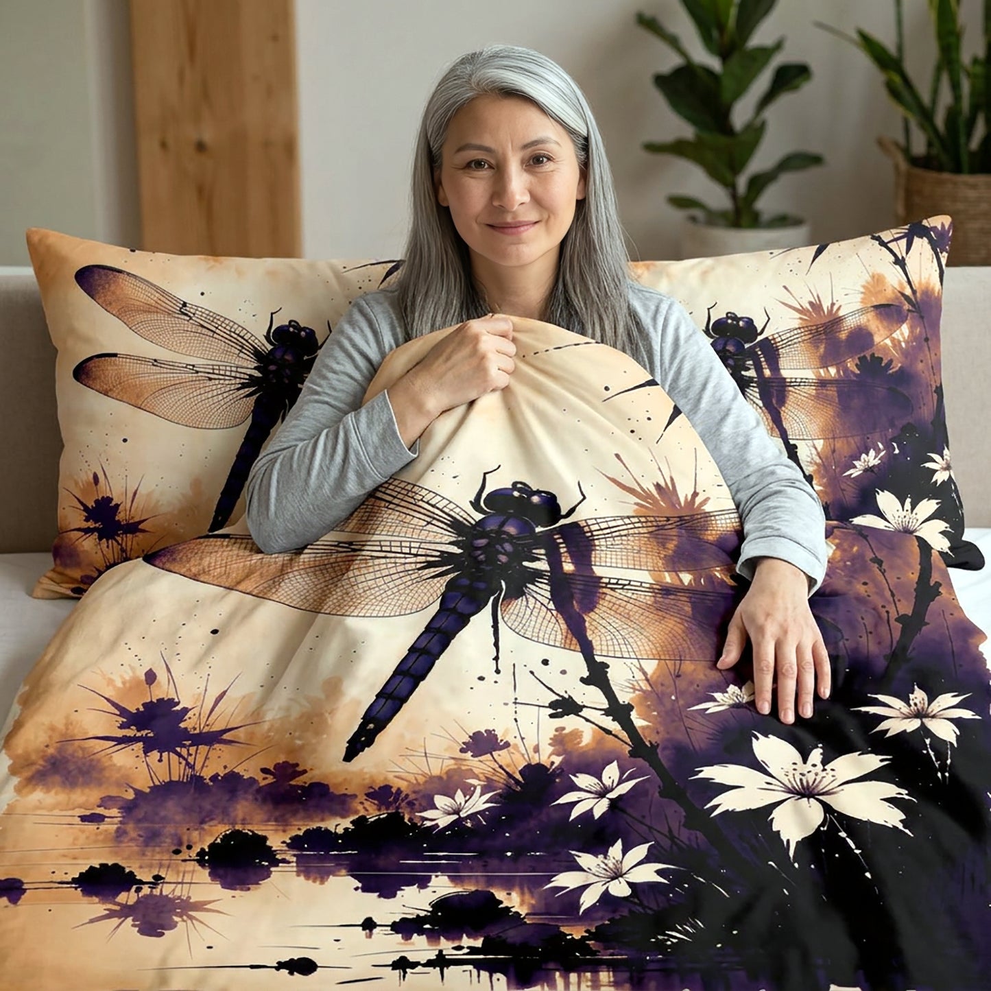 A woman with gray hair sits on a bed holding a duvet cover and matching pillowcase featuring a large dragonfly and floral watercolor design in beige, purple, black, and white.