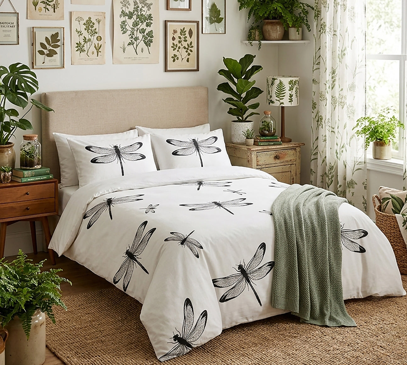 A neatly made bed in a styled bedroom featuring a white duvet cover and matching pillowcases printed with large black dragonfly motifs. The room includes a beige upholstered headboard, wooden nightstand, wall art, potted plants, sheer curtains, and a green throw blanket.