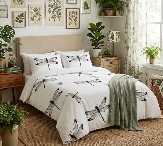 A neatly made bed in a styled bedroom featuring a white duvet cover and matching pillowcases printed with large black dragonfly motifs. The room includes a beige upholstered headboard, wooden nightstand, wall art, potted plants, sheer curtains, and a green throw blanket.