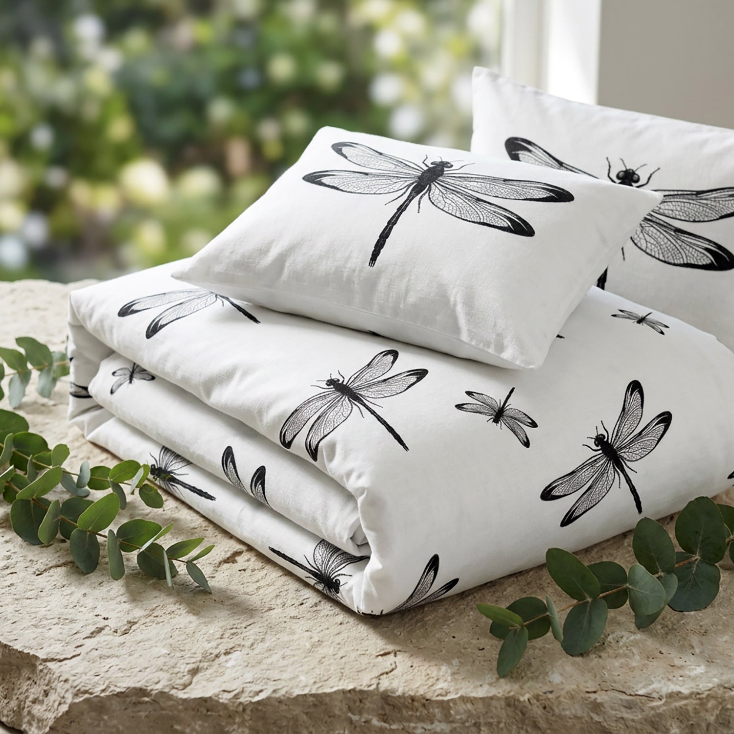 White bedding set with black dragonfly prints, including a folded duvet cover and matching pillowcases, styled on a stone surface with green eucalyptus leaves and a blurred garden background.
