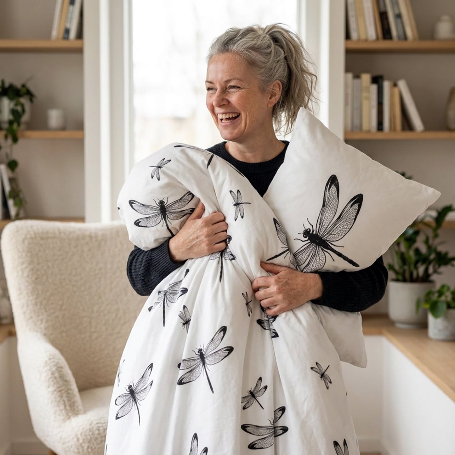 A woman in a cozy living room setting holds a white duvet cover and matching pillowcase printed with black dragonfly designs. The duvet cover features a subtle grid pattern on the reverse side.