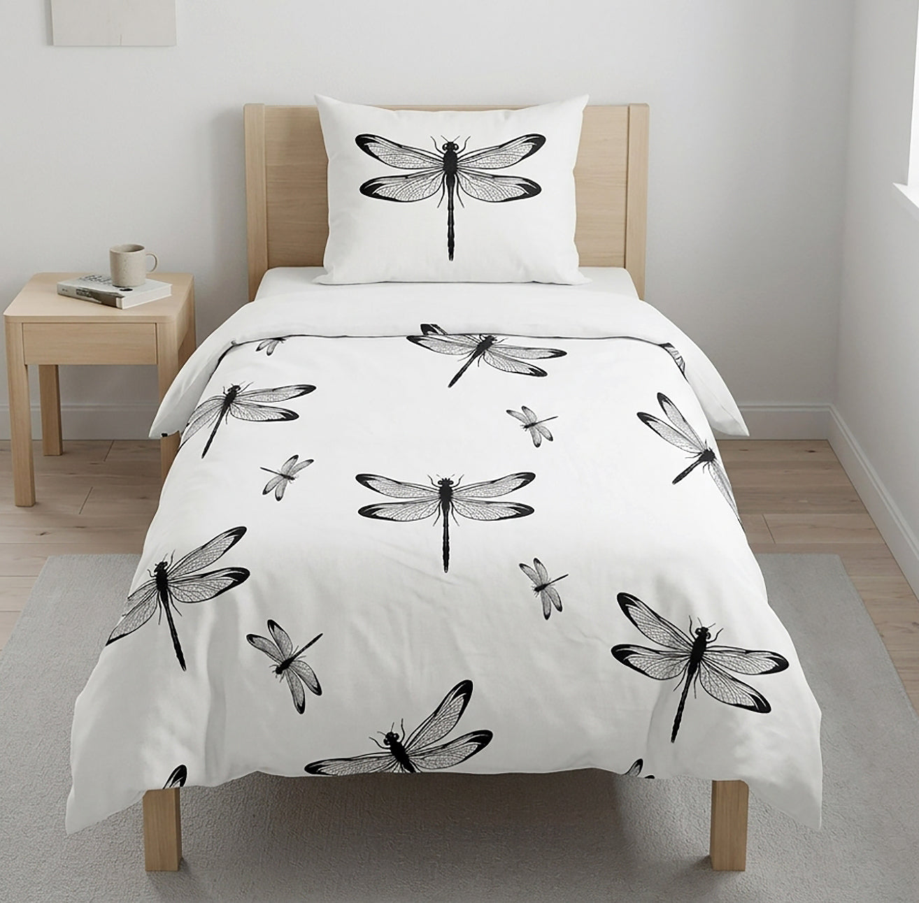 A single bed dressed in a white duvet cover and matching pillowcase, both printed with black dragonfly illustrations, shown in a bright bedroom with a light wood headboard, side table, and pale walls.