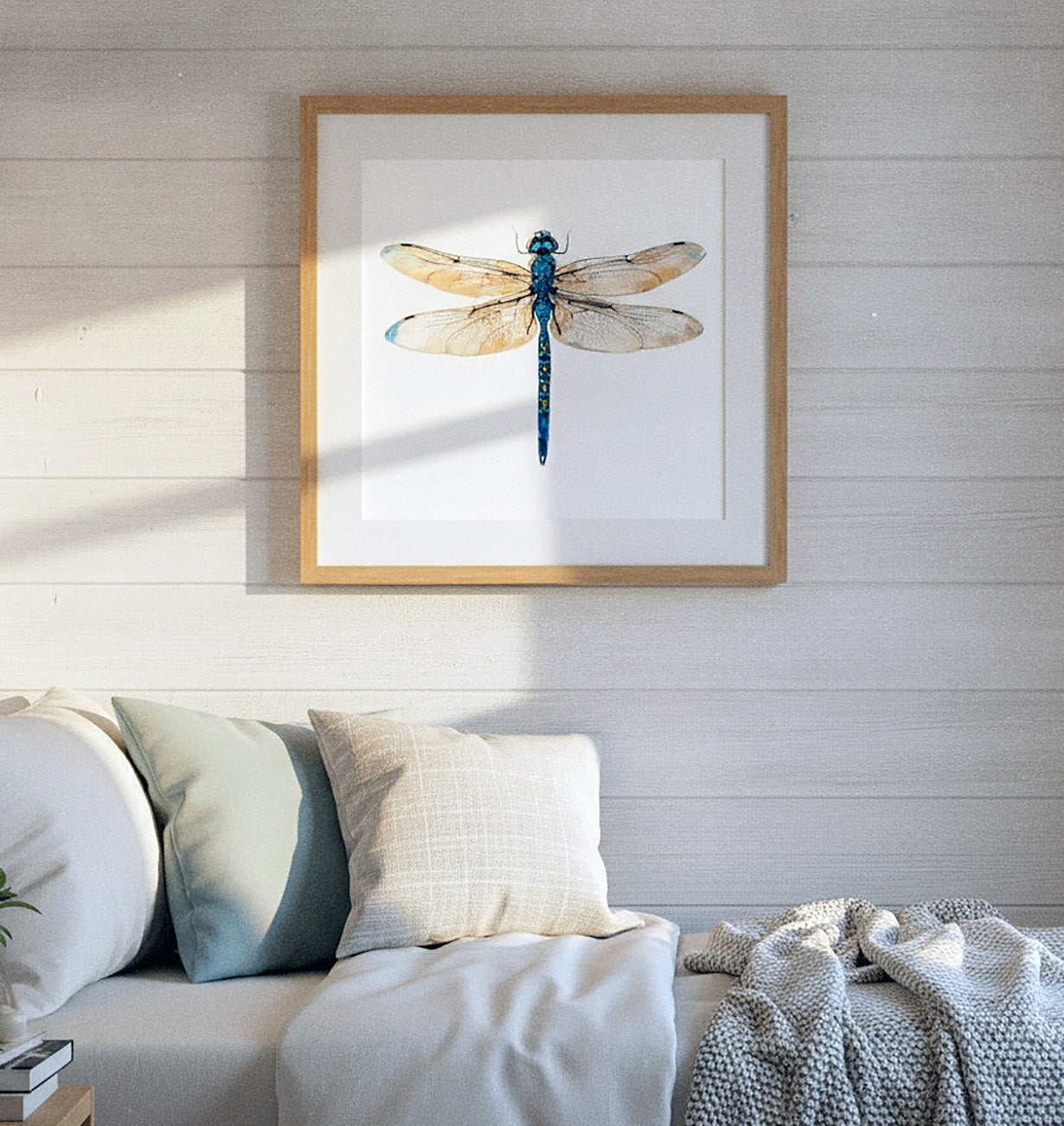 A framed watercolor dragonfly print hangs on a white shiplap wall above a bed with white and blue pillows and a gray knit blanket.