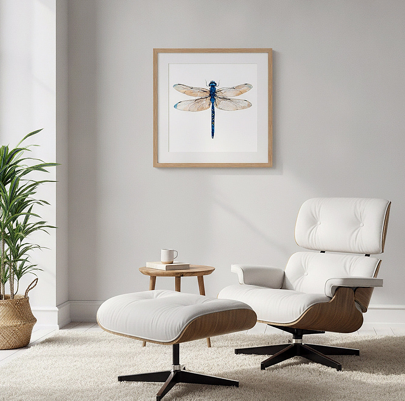 A framed dragonfly art print hangs on a light gray wall above a white lounge chair and matching ottoman in a modern living room setting.