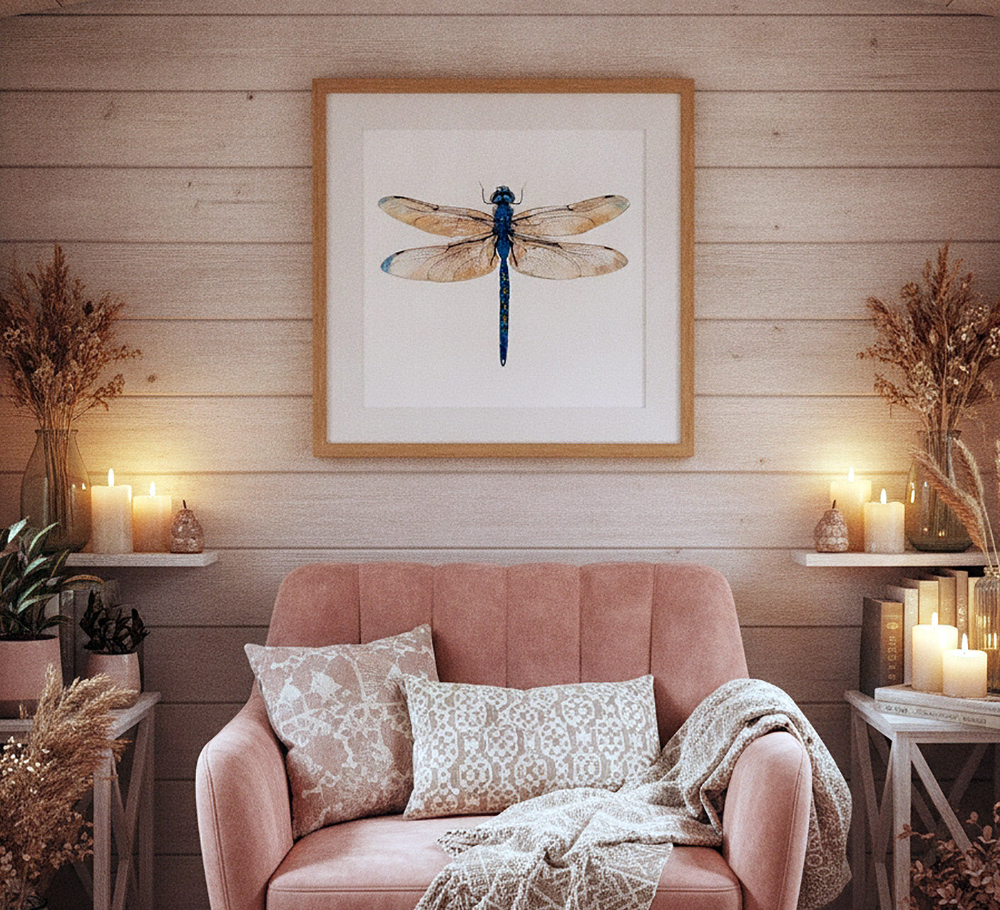 Framed wall art featuring a blue dragonfly on a white background, hung above a pink armchair in a styled room with candles, dried plants, and wood paneling.