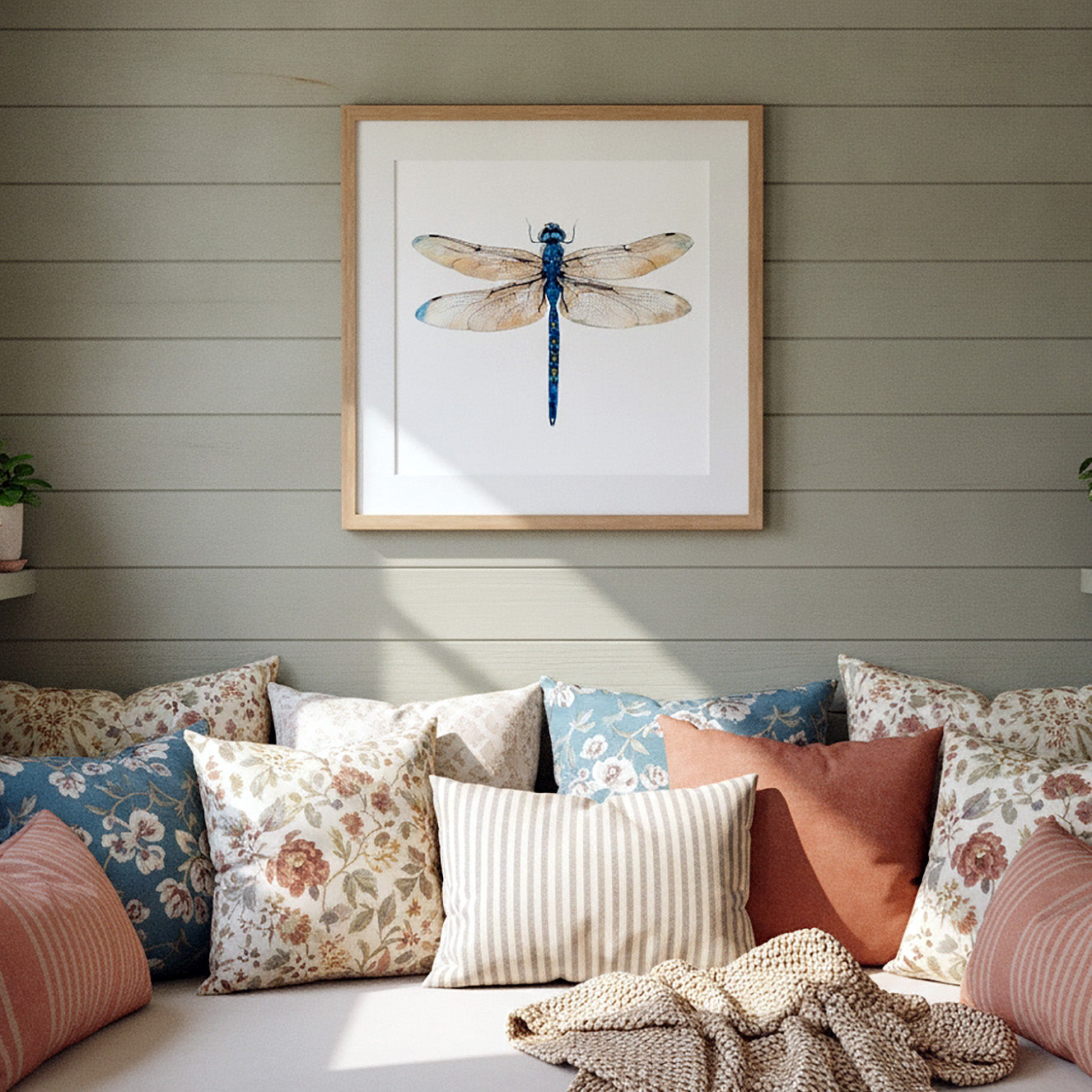 Framed wall art featuring a blue dragonfly on a white background, hung above a sofa with patterned pillows in a styled living room setting.