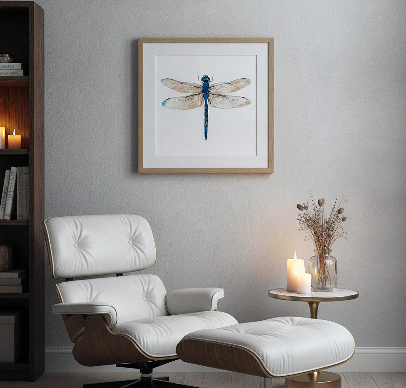 A framed watercolor dragonfly art print hangs on a light gray wall above a white leather lounge chair and matching ottoman, with a small side table and candles nearby.
