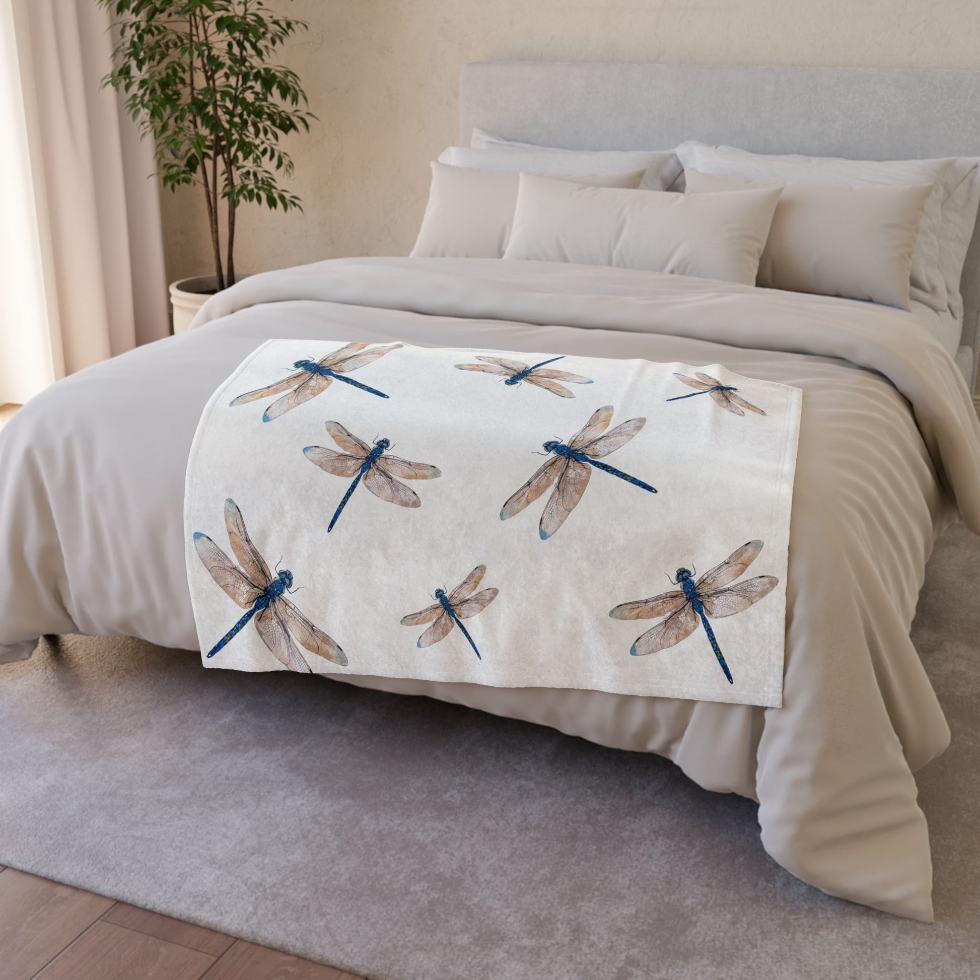 A white throw blanket draped over a bed, featuring a pattern of blue and brown dragonflies, set in a neutral bedroom environment.